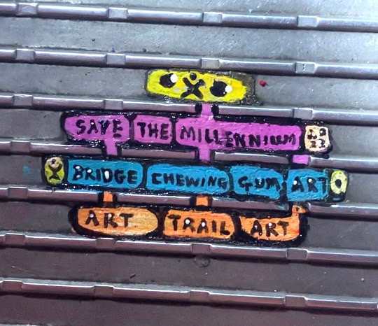 A yellow, purple, blue and orange slogan painting which says "save the millennium bridge chewing gum art trail art". 