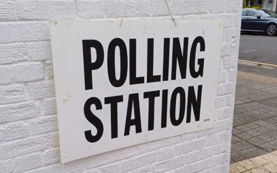 A polling station sign which is hung on a white wall.