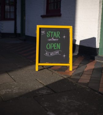 A sign on the street in the sun reading 'The Star of Greenwich open all welcome' with a yellow frame.
