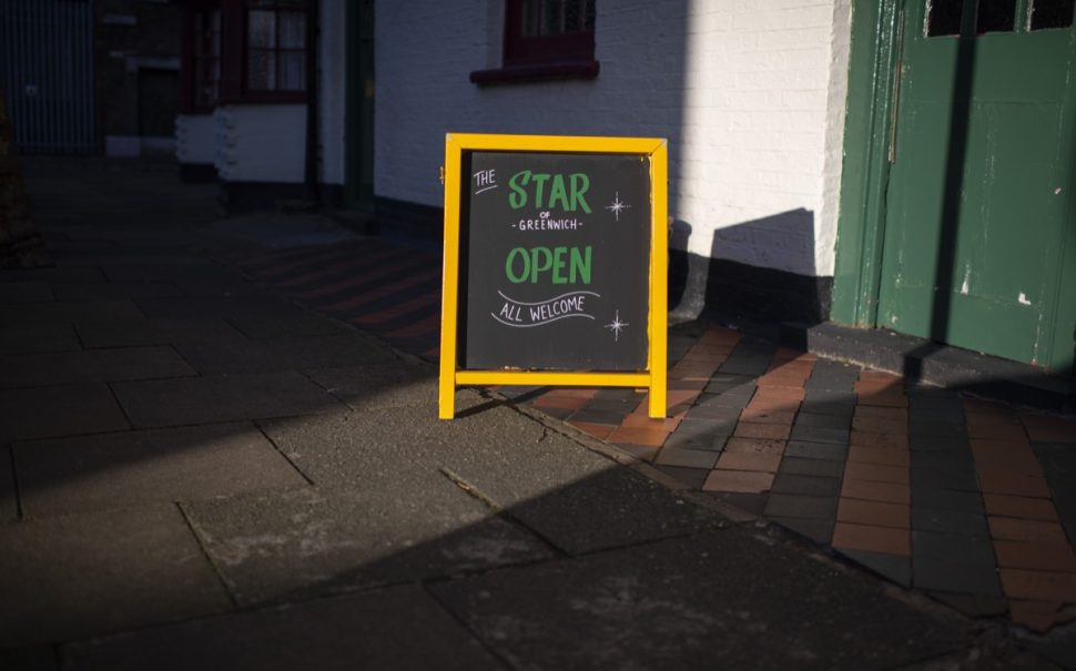 A sign on the street in the sun reading 'The Star of Greenwich open all welcome' with a yellow frame.