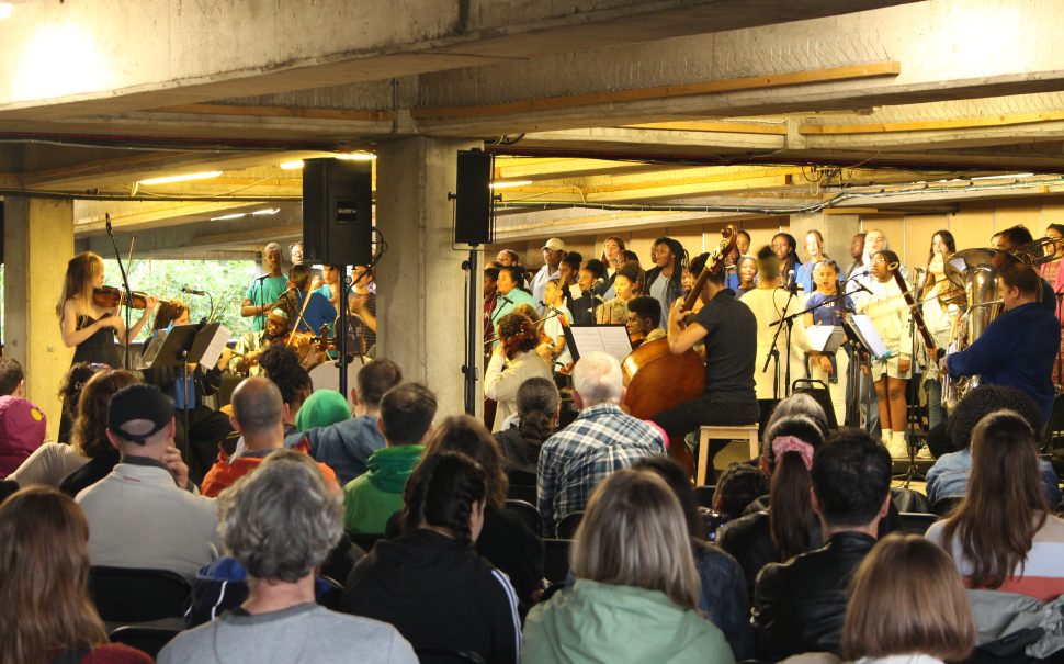 A seated crowd observe an orchestra playing in a multi-story car park