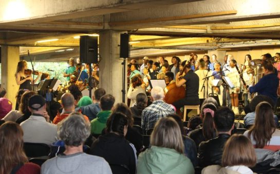 A seated crowd observe an orchestra playing in a multi-story car park