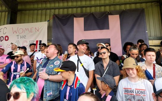 Fans of Dulwich Hamlet sing and watch the team with a pink and blue banner in the background at Champion Hill