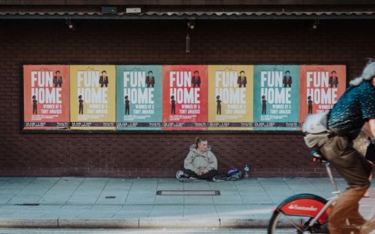 Photo shows a homeless women sitting on the street under posters of 'Fun Home'.