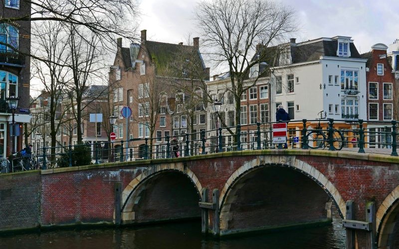 Photo of old brick bridge with three bows, crossing the canal water of the Prinsengracht in Amsterdam city downtown.