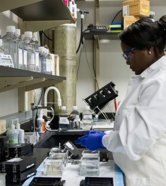 An image of a woman in a white lab technician coat standing over a sink in the lab.