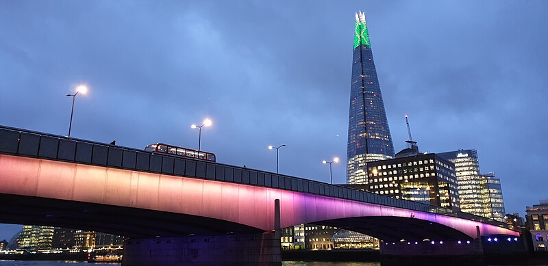 Picture of London Bridge, lit at night, from the lower, northern side of the river. The Shard is in the background to the upper-right.