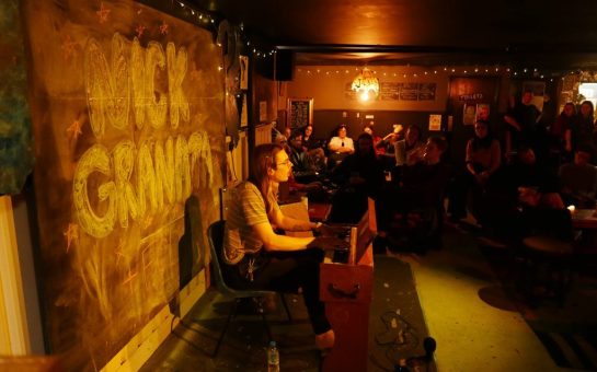 Image shows a musician playing the piano and singing, whilst people are crowded cosily round on sofas watching the performance.