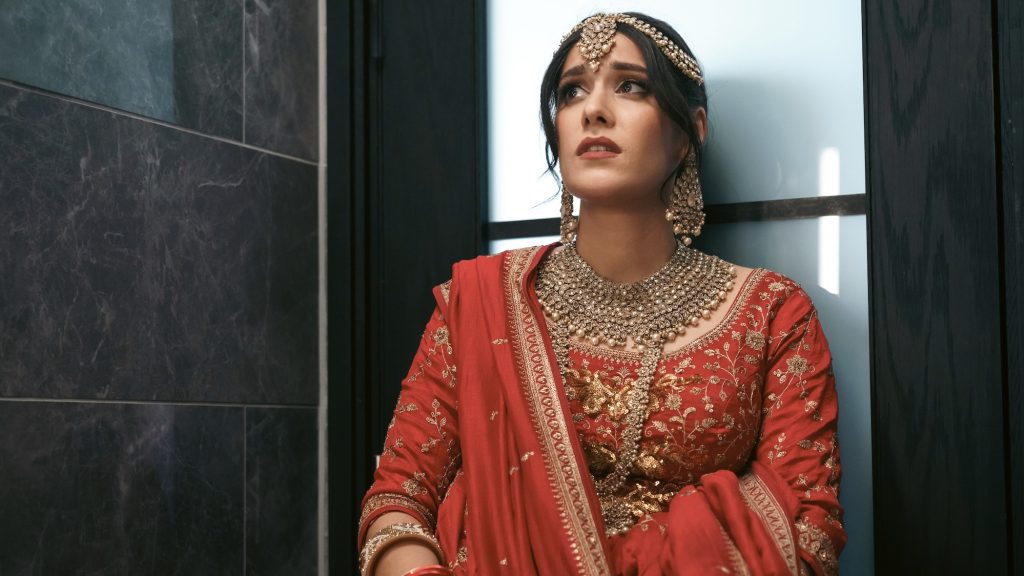  dressed in a traditional red and gold bridal outfit with intricate embroidery and heavy jewelry stands against a sleek, dark marble wall. She looks distressed, gazing upward with wide eyes, as if anxious or uncertain. Her ornate maang tikka (forehead ornament) and necklace glisten under the lighting.