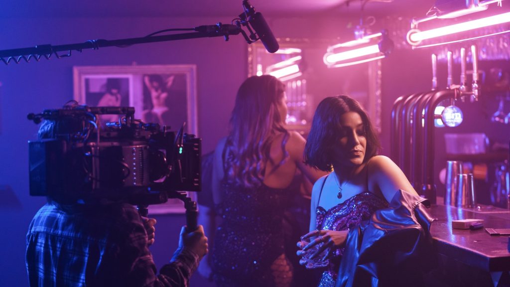 A behind-the-scenes shot of a film production in a dimly lit bar with neon pink and purple lighting. A woman with short dark hair, wearing a sequined dress and a leather jacket draped over her shoulders, leans on the bar with a drink in her hand. A camera operator films her, with a boom microphone visible above. In the background, another woman in a sparkly dress stands near a mirror, and framed photographs are on the wall.